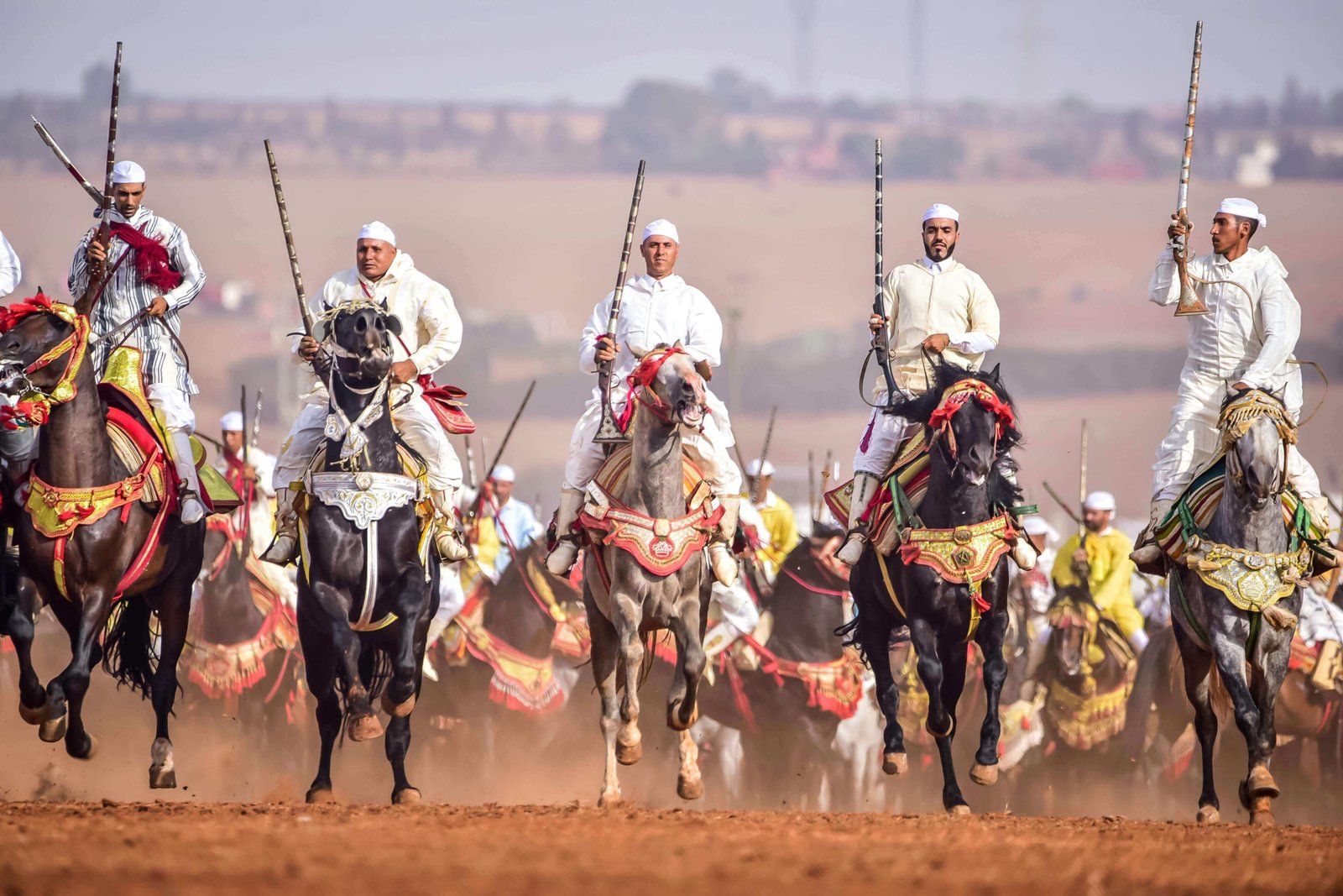 The-image-depicts-a-vibrant-traditional-Moroccan-fantasia-performance,-showcasing-skilled-riders-dressed-in-flowing-white-robes-atop-majestic-horses,-surrounded-by-a-backdrop-of-the-rich-cultural-heritage-of-Morocco.-.jpg