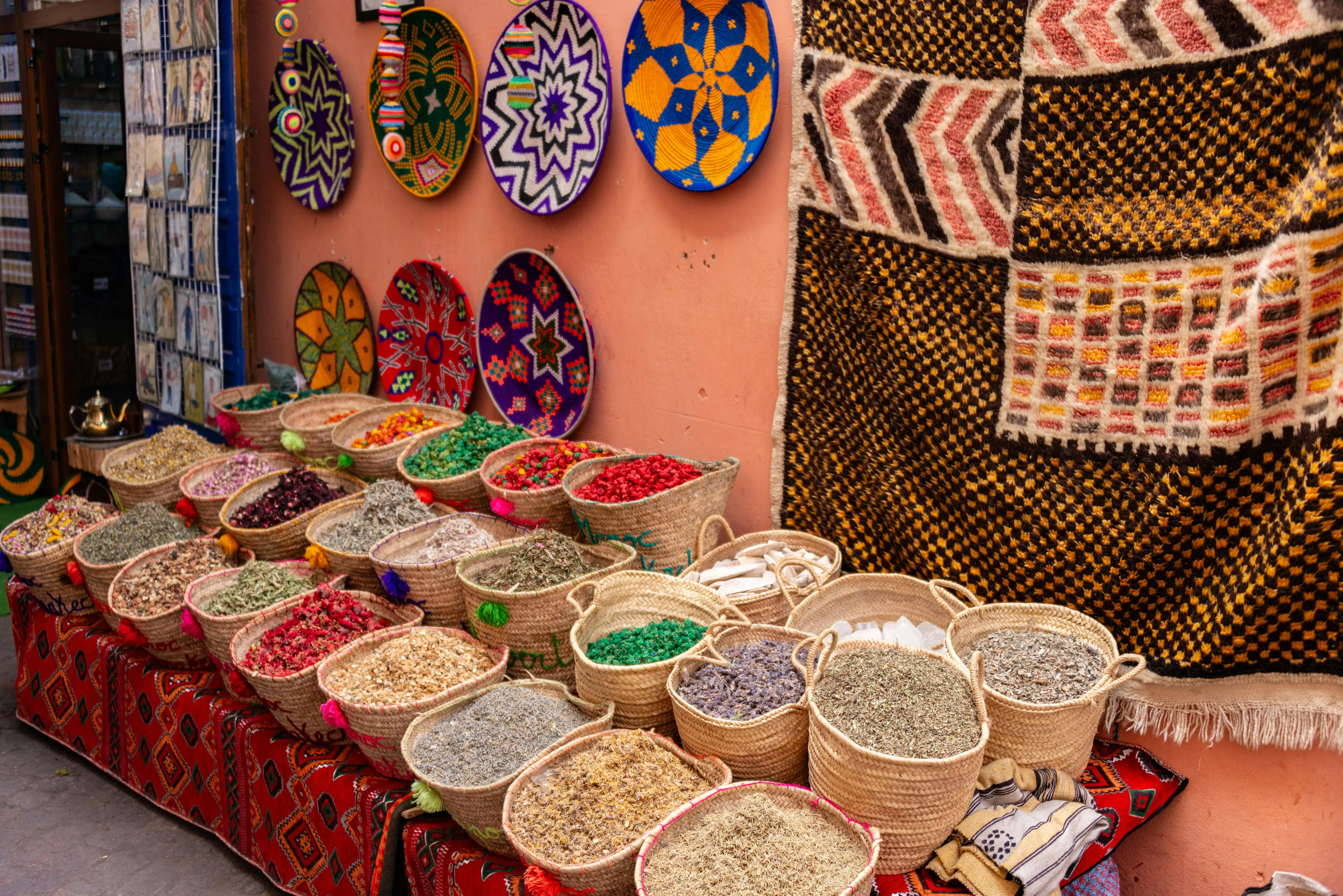 The-image-depicts-a-vibrant-traditional-Moroccan-market-scene,-showcasing-an-array-of-colorful-spices-and-intricate-textiles-displayed-in-bustling-souks.-This-lively-atmosphere-reflects-the-rich-local.webp